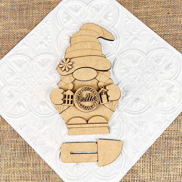 Enhance your home's farmhouse charm with Janet's Craft Corner's "Farmhouse Hello Gnome Shelf Sitter." This wooden cutout gnome features a hat, braids, and boots, holding a circle that reads "hello," all set against a textured white and beige background. Ideal as a DIY home décor craft kit for enthusiasts.