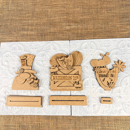 The Watermelon Mini Shelf Sitters DIY kit from Janet's Craft Corner includes three wooden cutouts: a top hat with "Home Sweet Home," a watermelon slice on a crate labeled "WATERMELON 25¢," and a flower pot with "One in a melon." Each farmhouse decor piece has its own stand.
