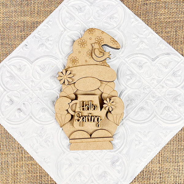 The Hellp Spring Girl Gnome Shelf Sitter by Janet's Craft Corner, a DIY seasonal home decor craft kit, features a wooden girl gnome with a floral hat holding a "Hello Spring" mug. It's adorned with flowers and a small bird against a textured white background.