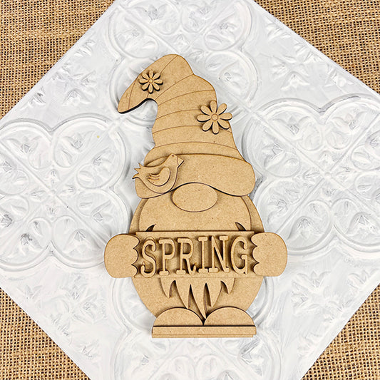 The Spring Sign Gnome Shelf Sitter by Janet's Craft Corner is a delightful DIY seasonal home decor kit. It includes a charming wooden gnome with a flower-detailed hat and bird, holding a "SPRING" sign against burlap with an embossed white pattern, perfect for enhancing your home.