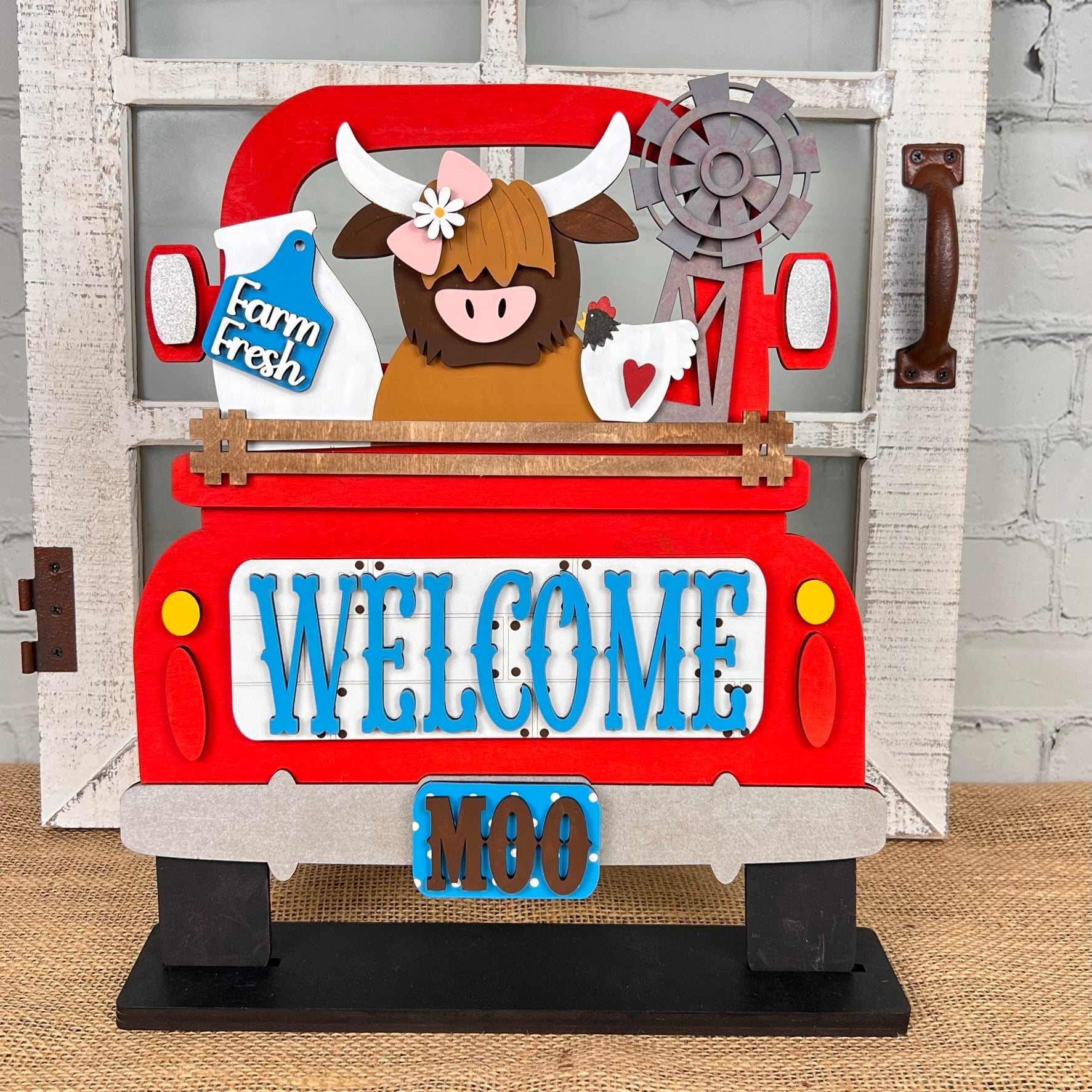 Discover a charming DIY home decor kit from Janet's Craft Corner: the Antique Truck Base with Interchangeable Insert. This decorative sign includes an antique truck scene with a brown cow, sunflowers, and a milk can in the back. The truck's tailgate is adorned with "WELCOME" in blue letters, accompanied by "MOO" beneath it. Complete with a rustic window and brick wall background, this set is perfect for adding interchangeable inserts to suit your style.