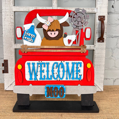 Discover a charming DIY home decor kit from Janet's Craft Corner: the Antique Truck Base with Interchangeable Insert. This decorative sign includes an antique truck scene with a brown cow, sunflowers, and a milk can in the back. The truck's tailgate is adorned with "WELCOME" in blue letters, accompanied by "MOO" beneath it. Complete with a rustic window and brick wall background, this set is perfect for adding interchangeable inserts to suit your style.