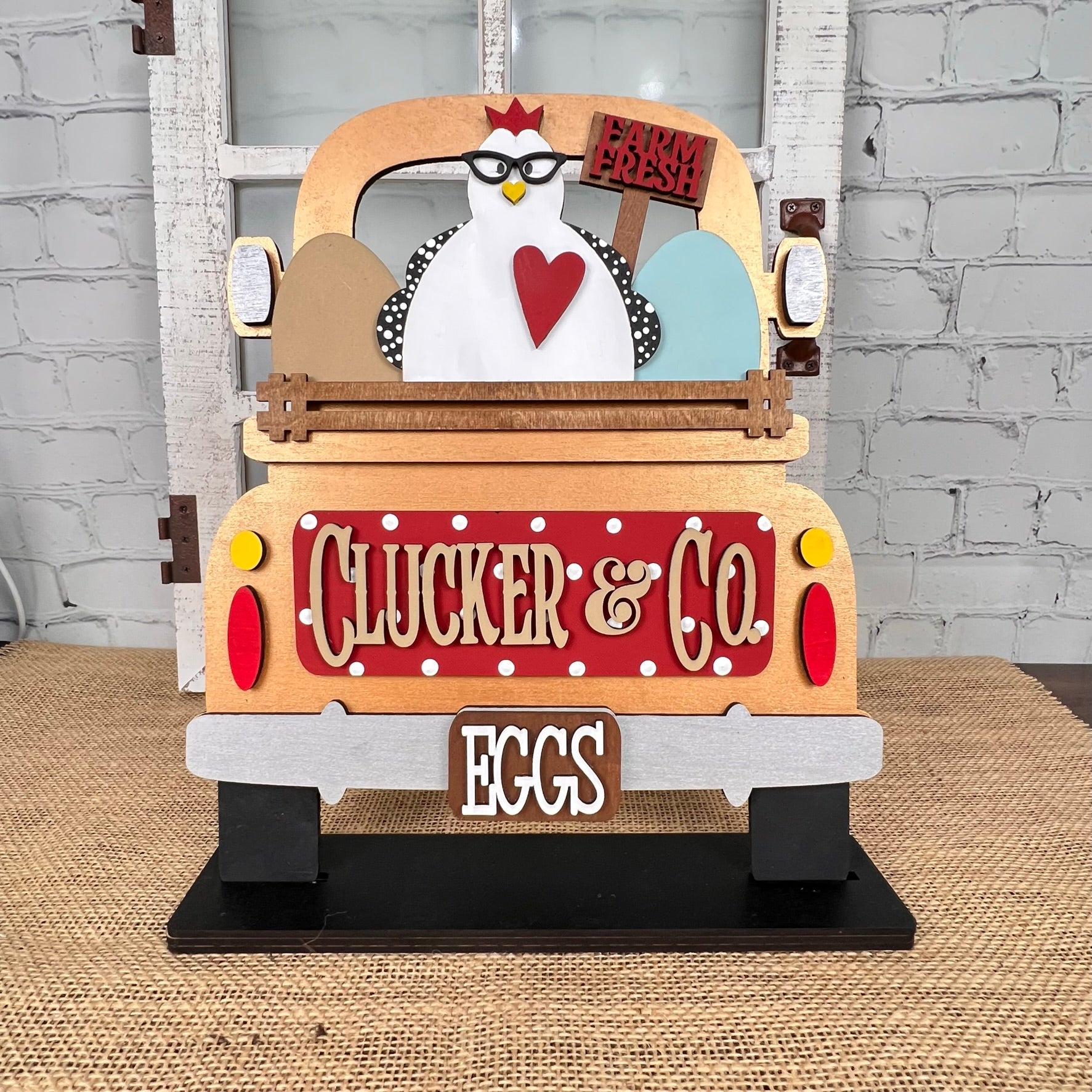 A rustic DIY home decor kit from Janet's Craft Corner showcases a chicken atop an antique truck base labeled "Clucker & Co. Eggs," holding a "Farm Fresh" sign, with three eggs nestled in the back, all set against a textured surface and brick wall. Ideal for fans of hand-painted crafts and rustic charm.