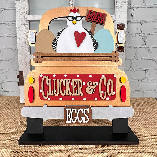 A decorative sign from Janet's Craft Corner, the Clucker & Co. Interchangeable Insert - Painted, features a vintage truck design with a charming chicken motif sporting glasses. The truck is adorned with the "Clucker & Co." and "EGGS" labels, highlighted by a heart symbol and three large eggs beneath a "Farm Fresh" banner, adding rustic charm to any space. This piece elegantly brings interchangeable craft inserts to life.