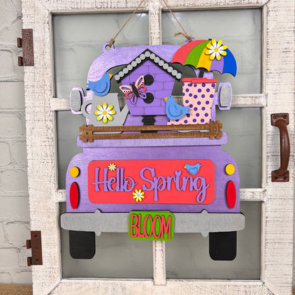 Introducing Janet's Craft Corner's DIY Home Decor Kit: an Antique Truck Base with Interchangeable Insert, perfect for welcoming spring. This vibrant design includes a "Hello Spring" sign adorned with bright purple, pink, and green colors. It features delightful hand-painted elements like flowers, birds, a butterfly, an umbrella, and a polka dot watering can. Ideal for crafting enthusiasts who enjoy creating their own decorative door hangings.