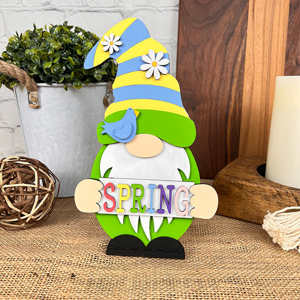 The "Spring Sign Gnome" from Janet's Craft Corner enhances your home décor with its vibrant hues. This colorful wooden figurine features a long white beard and a tall hat adorned with daisies and a blue bird. Holding a "SPRING" sign, it stands amongst plants and decorative items on a woven surface, embodying the spirit of the season.