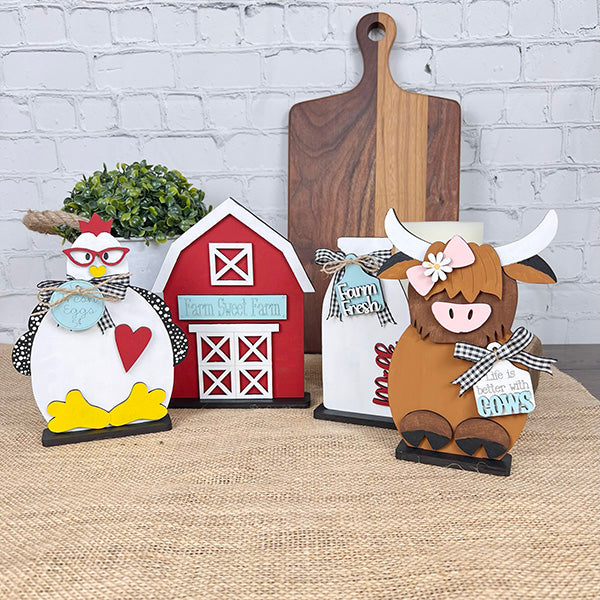 The Farm Standing Shelf Sitter from Janet's Craft Corner brings a rustic farmhouse scene to life with its charming DIY kit appeal, featuring a chicken, a red barn, a cow, and a "Farm Fresh" sign. Displayed against a brick wall and wooden cutting board on a burlap-covered surface, these decor items infuse any space with rustic charm.