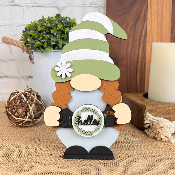 Introducing the "Farmhouse Hello Gnome" from Janet's Craft Corner, a charming figurine featuring red braids and a green and white striped hat. Holding a small sign with the word "hello," this gnome perfectly complements any farmhouse-style home décor. It is displayed on a burlap surface with a wooden backdrop, accompanied by a small plant and rustic accents.