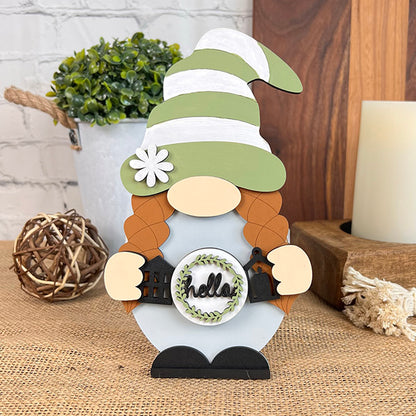 Introducing the "Farmhouse Hello Gnome" from Janet's Craft Corner, a charming figurine featuring red braids and a green and white striped hat. Holding a small sign with the word "hello," this gnome perfectly complements any farmhouse-style home décor. It is displayed on a burlap surface with a wooden backdrop, accompanied by a small plant and rustic accents.