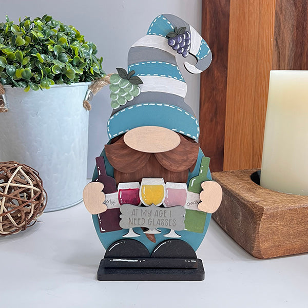 Add a touch of humor to your home with the Wine Lover Gnome from Janet's Craft Corner. Featuring a quirky design with a tall striped hat and holding three wine glasses, this charming gnome is complete with a sign that reads, "At my age I need glasses." Adorned with delightful grape accents, it's an ideal decorative piece for your table alongside a potted plant and candle.