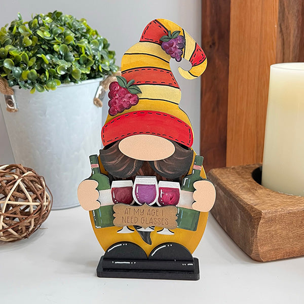 A charming Wine Lover Gnome by Janet's Craft Corner is decorated with a vibrant striped hat and grape accents, holding three wine glasses. The sign humorously states, "At my age, I need glasses." This delightful home décor piece is accompanied by a potted plant, a large candle, and a wicker ball.