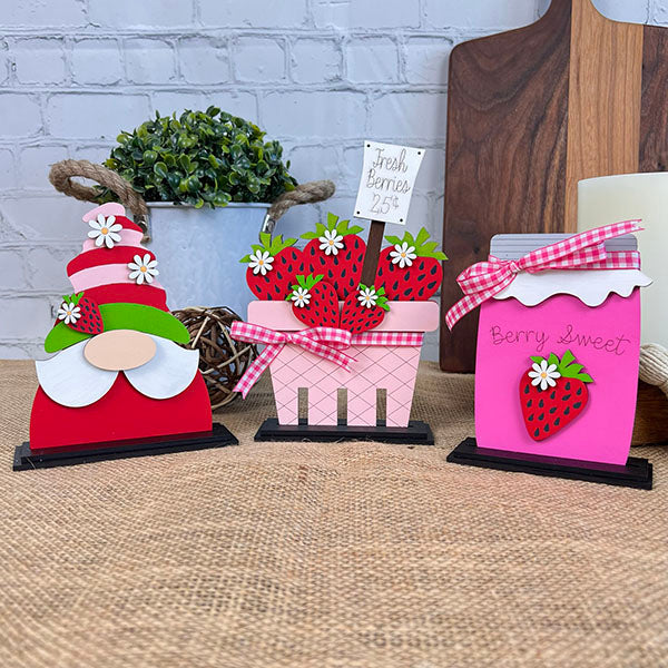 Displayed on a burlap table, the Strawberry Mini Shelf Sitters from Janet's Craft Corner, featuring a gnome with a strawberry hat, a pink basket labeled "Fresh Berries 25¢" filled with strawberries, and a pink jar labeled "Berry Sweet," each adorned with gingham ribbons and strawberry motifs, perfectly encapsulate the farmhouse chic vibe.