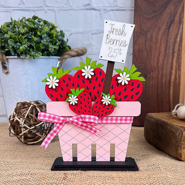 A decorative display showcases a pink basket filled with wooden red strawberries and small white flowers, complemented by Janet's Craft Corner's Strawberry Mini Shelf Sitters. A sign reading "Fresh Berries 25¢" adds charm. The basket is placed on a burlap-covered surface against a brick wall, exuding farmhouse chic appeal.