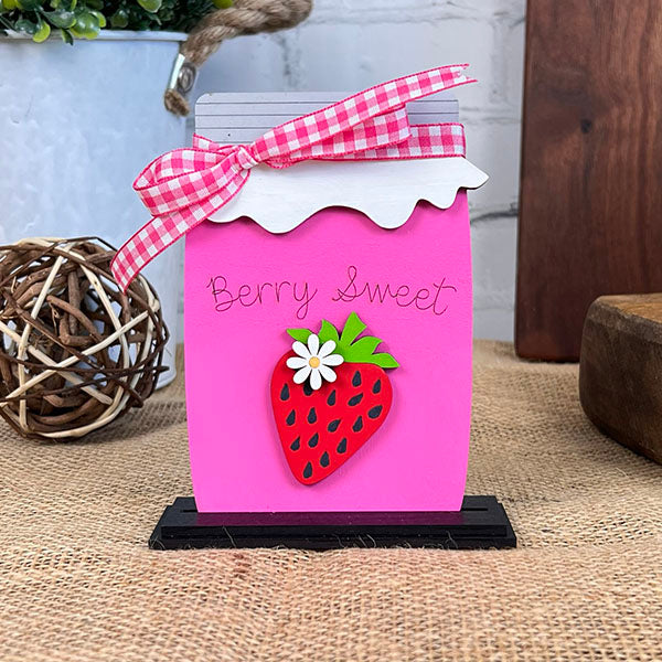 Introducing the "Strawberry Mini Shelf Sitters" by Janet's Craft Corner, a charming pink wooden block shaped like a jar. It is adorned with a delightful red strawberry and white flower on the front, adding a farmhouse chic touch to your space. The phrase "Berry Sweet" is elegantly displayed in cursive, complemented by a pink and white checkered ribbon on top. Perfect for enhancing your strawberry-themed decor with its wicker background design.