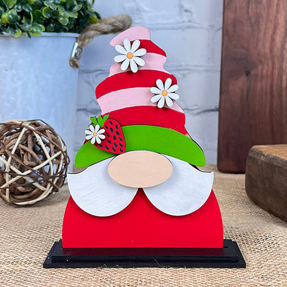 The Strawberry Mini Shelf Sitter from Janet's Craft Corner exemplifies farmhouse chic with its charming decor. This decorative wooden gnome features a striped pink and red hat adorned with white flowers and a strawberry. Its white beard and black base complement the rustic backdrop of a plant and twine ball, creating a delightful display.