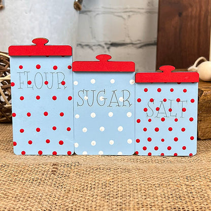 The "Baking - Tiered Tray Kit" from Janet's Craft Corner brings a charming touch to any farmhouse decor with its decorative kitchen canisters. Labeled "Flour," "Sugar," and "Salt," they feature blue backgrounds adorned with red and white polka dots, topped with red lids, all beautifully displayed on a rustic burlap surface.