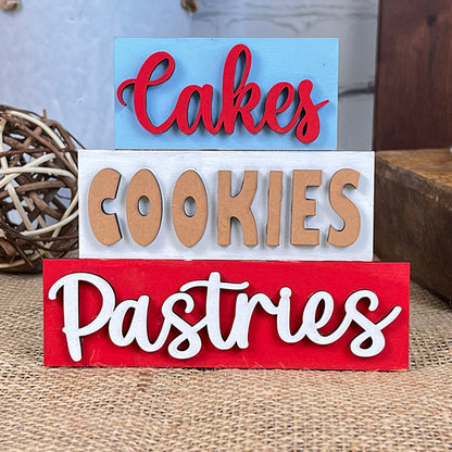 The Baking - Tiered Tray Kit by Janet's Craft Corner features stacked wooden signs placed on a burlap surface, ideal for adding a farmhouse decor touch. The signs display the words "Cakes" in red on blue, "Cookies" in beige on white, and "Pastries" in white on red. A woven ball sits in the background, enhancing the charm of this delightful kit.