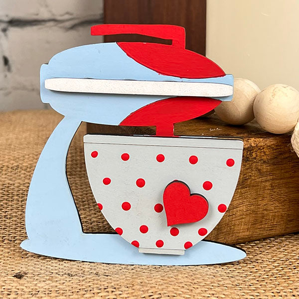 Introducing the Baking - Tiered Tray Kit from Janet's Craft Corner. This delightful DIY kit includes a decorative wooden figure shaped like a vintage stand mixer, beautifully painted in blue and red. A gray mixing bowl adorned with red polka dots and a heart adds extra charm to the piece, making it perfect for farmhouse decor. It complements any setting with its burlap backdrop, wooden block, and beads.