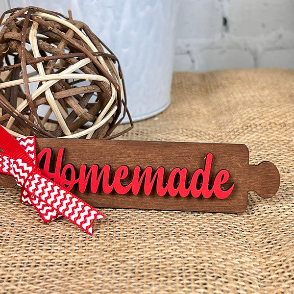 The "Baking - Tiered Tray Kit" from Janet's Craft Corner features a charming wooden sign with red text that reads "Homemade," ideal for farmhouse decor. It is beautifully adorned with a red and white chevron ribbon. The display also includes a woven decorative ball and a white pot set on a burlap surface, adding an extra touch of elegance to any arrangement.