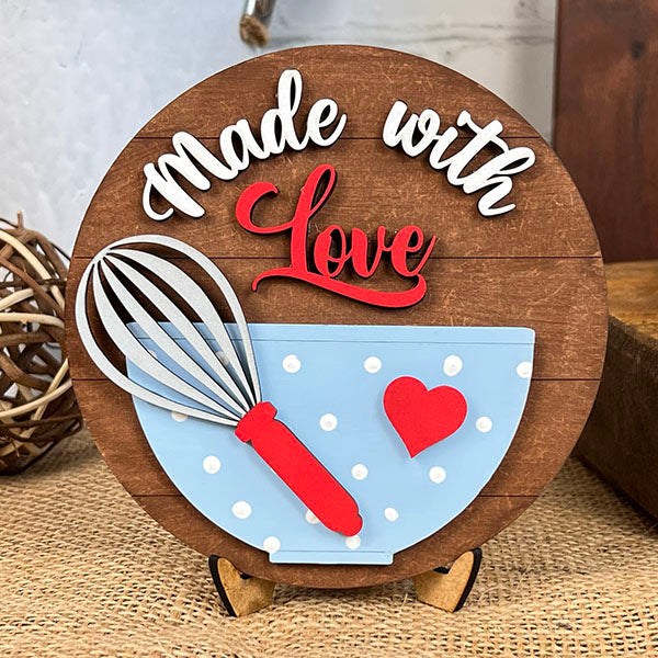 The "Baking - Tiered Tray Kit" by Janet's Craft Corner includes a charming decorative round wooden plaque that showcases the words "Made with Love" in elegant white and red cursive. Positioned below, a blue polka dot bowl adorned with a red heart and accompanied by a whisk rests on a rustic burlap surface.