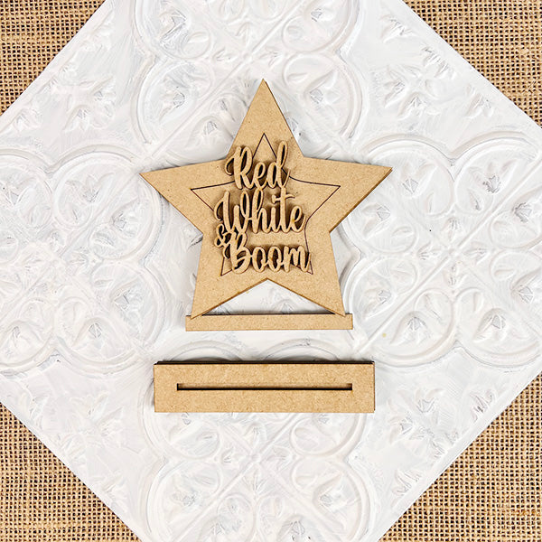 A wooden star with the phrase "Red, White & Boom" cut out at its center is set against a textured white backdrop. This Americana figure from Janet's Craft Corner, known as the Patriotic Shelf Sitters, includes a separate rectangular wooden base for support and makes an ideal patriotic addition to any shelf.