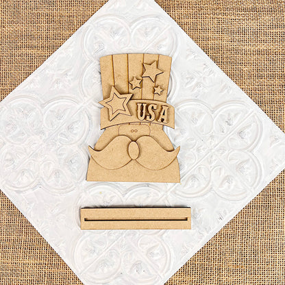 The Patriotic Shelf Sitters by Janet's Craft Corner spotlight an Americana figure resembling Uncle Sam's hat adorned with stars and the word "USA," showcasing a prominent mustache and resting on a slotted base against a textured white and burlap background.