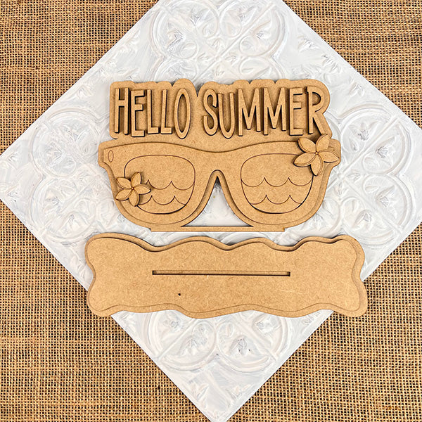 Wooden cutout sunglasses with "HELLO SUMMER" text and floral details are on a textured mat. Below, the Hello Summer Sunglasses Shelf Sitter by Janet's Craft Corner is visible, perfect for farmhouse decor enthusiasts. The background highlights a woven fabric design.