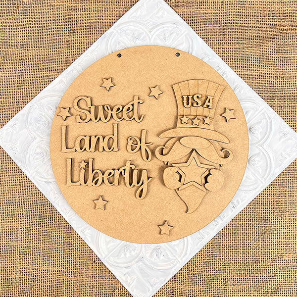 The Sweet Land of Liberty Door Hanger by Janet's Craft Corner features a round wooden plaque with "Sweet Land of Liberty" and a gnome in a USA hat. Stars accent the burlap and quilted white backdrop, making it perfect for patriotic decor enthusiasts. This DIY craft kit includes one set.