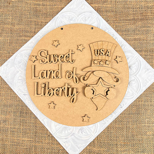 The Sweet Land of Liberty Door Hanger by Janet's Craft Corner features a round wooden plaque with "Sweet Land of Liberty" and a gnome in a USA hat. Stars accent the burlap and quilted white backdrop, making it perfect for patriotic decor enthusiasts. This DIY craft kit includes one set.