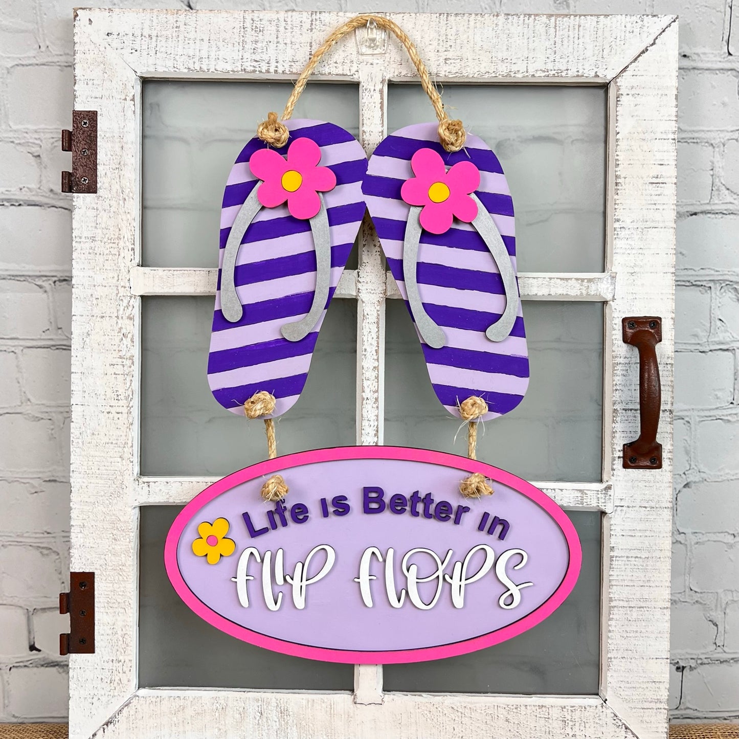 On a white framed glass door, a decorative Flip Flops Door Hanger by Janet's Craft Corner features purple and white striped flip-flops adorned with pink flowers. Below, a pink oval plaque joyfully declares, "Life is Better in Flip Flops," bringing a charming touch to your home décor.