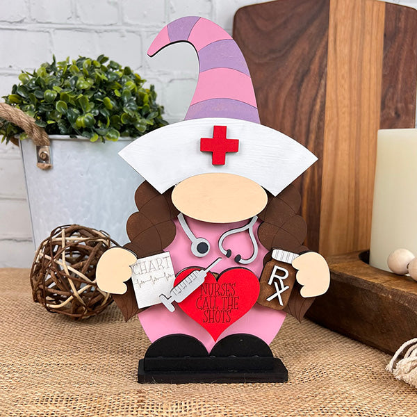 Introducing the Nurse Gnome from Janet's Craft Corner, a delightful wooden figurine featuring a pink hat and a stethoscope. This DIY kit also comes with a chart and syringe accessory. The gnome holds a red heart inscribed with "Nurses Call the Shots," making it an ideal addition to farmhouse decor. Perfectly suited for display on tabletops surrounded by decorative plants and wooden accents.