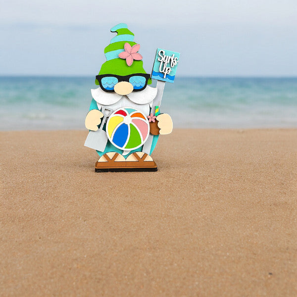 Janet's Craft Corner offers the Beach Gnome Shelf Sitter DIY home decor craft kit, featuring a whimsical gnome on a sandy beach with sunglasses, a green hat adorned with a pink flower, and holding a "Surf's Up" sign and beach ball against an ocean backdrop.