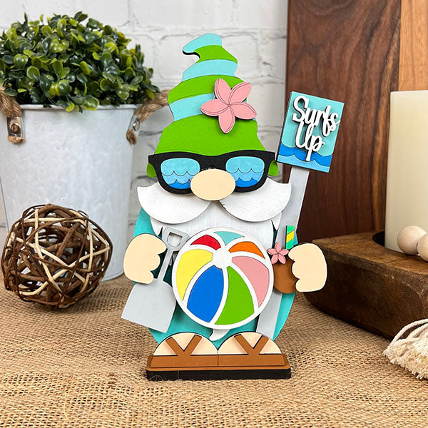 The Beach Gnome from Janet's Craft Corner is part of a delightful beach decor collection, featuring sunglasses and a green hat while holding a "Surf's Up" sign. It stands beside a colorful beach ball and a tropical drink. The setting includes a plant in a white pot and decorative items on wooden surface.