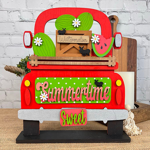 The Summertime Watermelon Interchangeable Insert - Painted, by Janet's Craft Corner, showcases a charming red truck with hand-painted ornaments of watermelons and daisies in the back. The tailgate is adorned with the words "Summertime" and "Sweet." An accompanying small wooden crate labeled "Watermelons," surrounded by polka dots and black cat silhouettes, adds interchangeable home decor charm.