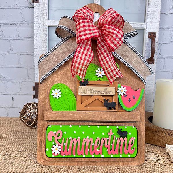 Introducing the **Summertime Watermelon Interchangeable Insert - Painted** by *Janet's Craft Corner*: This hand-painted decoration showcases a red checkered bow and a watermelon slice, paired with a green "Summertime" sign accented with white dots. Enhanced with black silhouettes of cows and white daisy flowers, this charming piece is perfect for your home decor.