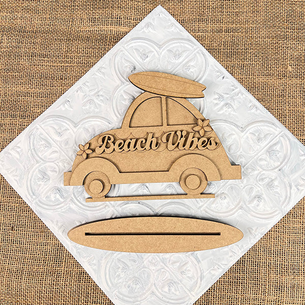 The Beach Vibes Volkswagen Shelf Sitter by Janet's Craft Corner is a DIY home decor kit. Shaped like a vintage car with a surfboard, floral details, and "Beach Vibes" inscription, it complements textured white or burlap backgrounds.