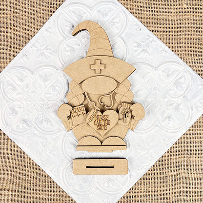 The Nurse Gnome Shelf Sitter from Janet's Craft Corner is a DIY home decor craft kit featuring a wooden cutout shaped like a nurse gnome with medical items and "Nurses Call the Shots" text, perfect for farmhouse decor against its patterned background.