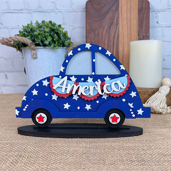 The America Volkswagen from Janet's Craft Corner is a charming Americana figure, featuring a decorative wooden car painted blue with white stars and "America" in bold red and white lettering. It rests on star-detailed red and white wheels, displayed on a burlap surface surrounded by home décor elements such as plants and a softly glowing candle.