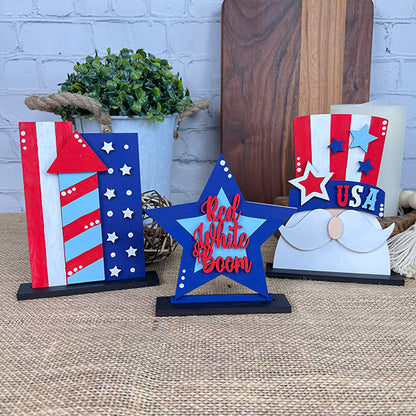 Explore Janet's Craft Corner's Patriotic Shelf Sitters, showcasing festive Americana figures in vibrant red, white, and blue tones. The collection includes wooden blocks shaped like a tie and shirt, a star adorned with "Red, White, and Boom," and a mustache perched on a striped block embossed with "USA." These delightful pieces are accompanied by greenery against a backdrop of wooden boards. Ideal for adding patriotic charm to your decor!