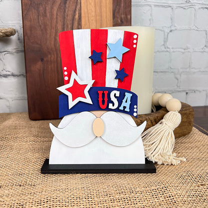 The Patriotic Shelf Sitters from Janet's Craft Corner showcase a decorative Americana figure wearing a red, white, and blue Uncle Sam hat with stars, the letters "USA," and a white mustache. It is elegantly displayed on a woven mat with a wooden tray and a candle in the background.