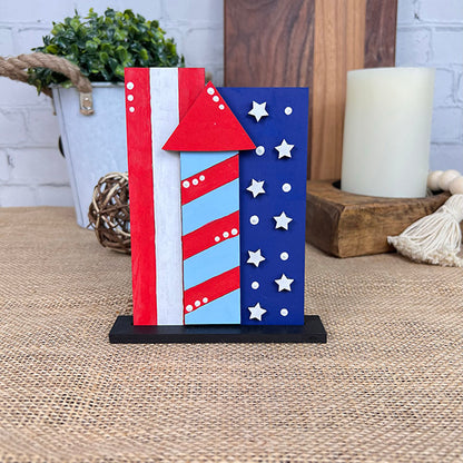 A decorative piece from Janet's Craft Corner, the Patriotic Shelf Sitters collection features a wooden block shaped like a firework, adorned with vibrant red, white, and blue stripes and stars. It stands upright on a black base and is showcased on a burlap surface beside a candle, plant, and wooden backdrop.