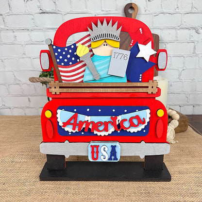 The America Interchangeable Insert DIY home decor kit by Janet's Craft Corner is a charming decorative red wooden truck adorned with a "USA" license plate and an "America" sign, embodying Americana spirit. It's filled with patriotic items such as a miniature Statue of Liberty, an American flag, and a star—ideal for crafting against the backdrop of a white brick wall.