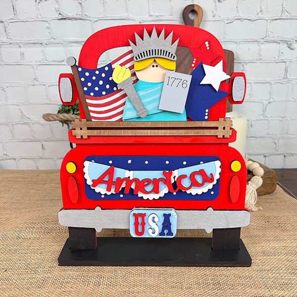 Introducing the Antique Truck Base with Interchangeable Insert DIY home decor kit from Janet's Craft Corner. This red antique truck model features American patriotic decorations, including a flag, a Statue of Liberty figure, and a "1776" sign. The back displays "America" with "USA" beneath it, positioned against a light brick background—ideal for interchangeable inserts or as part of your hand-painted DIY craft projects.
