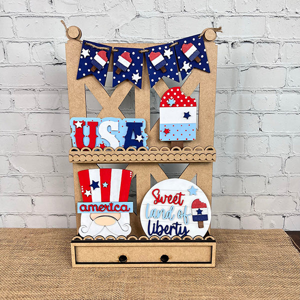 A decorative wooden shelf exudes farmhouse-style decor, featuring various patriotic items such as a "Sweet Land of Liberty" sign, USA letters, and flag-themed art. Above the display hangs a bunting adorned with American flag patterns, creating the perfect showcase for your Patriotic - Tiered Tray Kit from Janet's Craft Corner against a white brick wall.