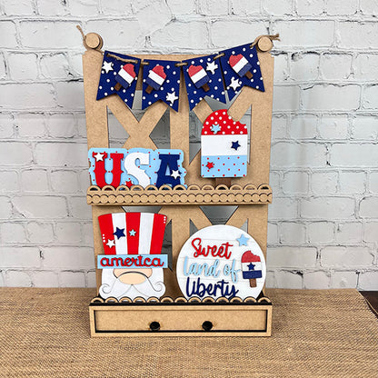 A decorative wooden shelf exudes farmhouse-style decor, featuring various patriotic items such as a "Sweet Land of Liberty" sign, USA letters, and flag-themed art. Above the display hangs a bunting adorned with American flag patterns, creating the perfect showcase for your Patriotic - Tiered Tray Kit from Janet's Craft Corner against a white brick wall.