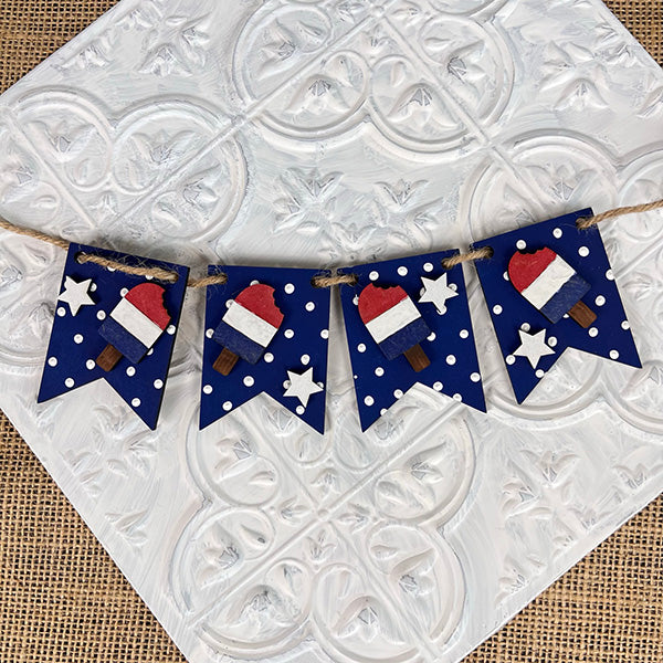 A decorative banner featuring four flags is set against a textured white and burlap backdrop, ideal for farmhouse-style decor. Each flag is navy blue adorned with white polka dots, showcasing a painted ice cream pop in red, white, and blue layers with star accents. Perfect for the Patriotic - Tiered Tray Kit by Janet's Craft Corner.