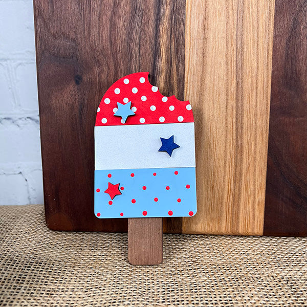 This wooden decorative popsicle from Janet's Craft Corner showcases a patriotic red, white, and blue design. The top is adorned with red polka dots and stars, the middle section is a crisp white, and the bottom sparkles in blue with dots and stars. It's an ideal addition to the Patriotic - Tiered Tray Kit for a charming display against a rustic wooden background.