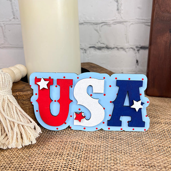 The image features a farmhouse-style decor sign from Janet's Craft Corner's "Patriotic - Tiered Tray Kit." The sign spells out "USA" in red, white, and blue with small stars and is arranged on burlap next to a candle and tassel against a white brick wall.