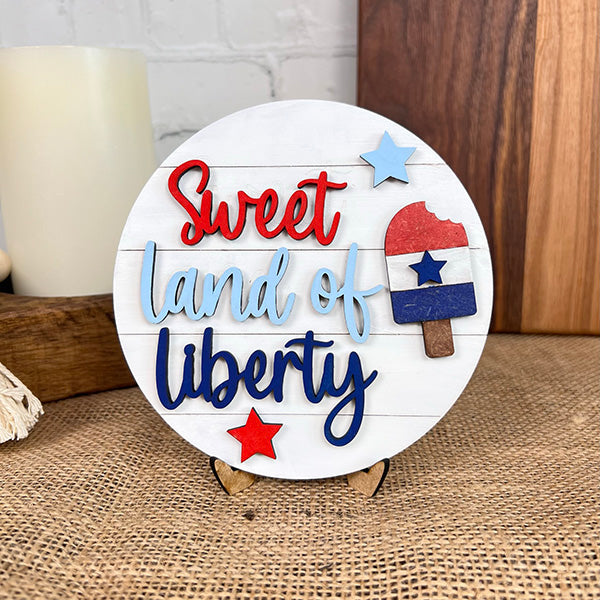 This farmhouse-style Patriotic - Tiered Tray Kit from Janet's Craft Corner showcases a round sign featuring "Sweet land of liberty" in red and blue on a white background. Accompanied by a red, white, and blue popsicle with a bite taken out to the right, the sign is adorned with red and blue stars. It is beautifully displayed on a small stand.