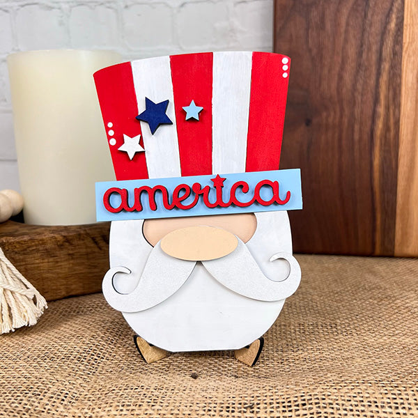 This farmhouse-style decor from Janet's Craft Corner showcases the Patriotic - Tiered Tray Kit, featuring a decorative wooden figure of a bearded man with a star-adorned red, white, and blue top hat emblazoned with "America" on the band. Ideal for enhancing your patriotic tiered tray, it is elegantly displayed on burlap alongside a candle and wooden boards in the background.